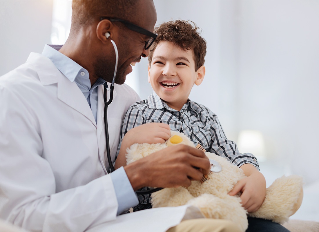 Employee Benefits - Portrait of a Cheerful Young Male Doctor with a Young Boy Giving a Teddy Bear a Medical Exam in an Office