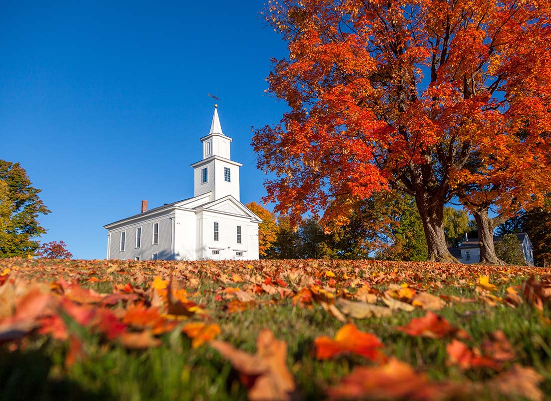 Insurance by Industry - Landscape View of a White Church Against a Bright Blue Sky with Colorful Fall Trees and Leaves Fallen on the Grass