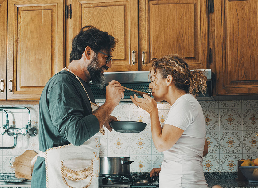 Personal Insurance - Cheerful Middle Aged Husband Wearing an Apron Letting his Wife Try the Food he Cooked as They Stand in the Kitchen Together